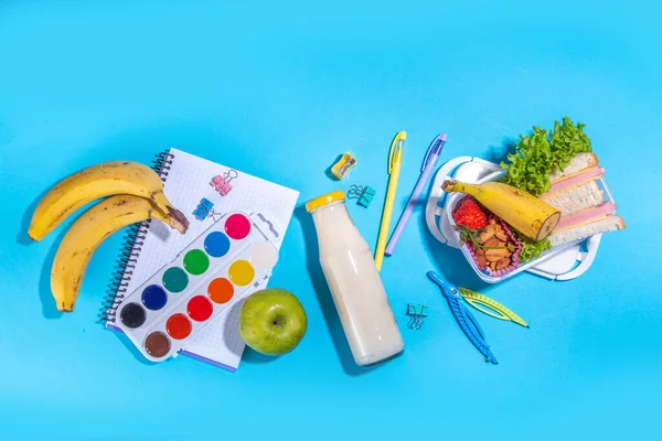 Sağlıklı okul beslenme çantası: sandviç, sebze, meyve, fındık ve yoğurt, içinde okul çocukları malzemeleri, aksesuarlar ve yüksek renkli mavi arka plan fotokopi alanı üzerine sırt çantası