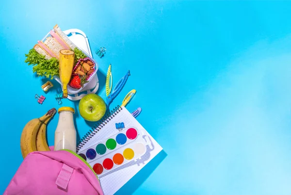 Sağlıklı okul beslenme çantası: sandviç, sebze, meyve, fındık ve yoğurt, içinde okul çocukları malzemeleri, aksesuarlar ve yüksek renkli mavi arka plan fotokopi alanı üzerine sırt çantası