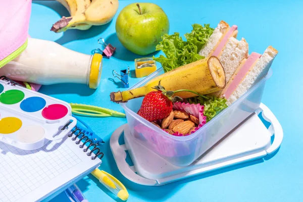 Sağlıklı okul beslenme çantası: sandviç, sebze, meyve, fındık ve yoğurt, içinde okul çocukları malzemeleri, aksesuarlar ve yüksek renkli mavi arka plan fotokopi alanı üzerine sırt çantası