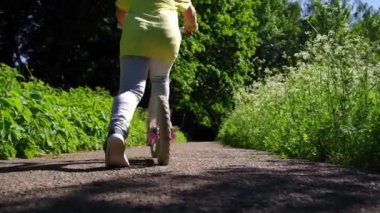 Yazın açık hava aktiviteleri videosu. Çocukların bacaklarına yakın çekim ve parkta bir scooter, kız itiyor ve scooterla ileri doğru gidiyor. Küçük kız güneşli bir günde scooter kullanıyor.