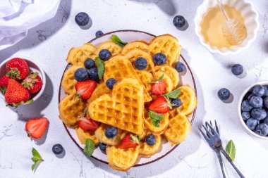 Çilekli, yaban mersinli ve ballı Belçika waffle 'ları, beyaz mutfak masalarında ev yapımı kalp şeklinde waffle' lar.