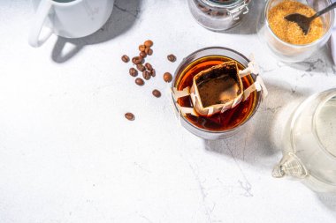 Modaya uygun demlenmiş damlatılmış kahve, soğuk demlenmiş kahve. Damlatma filtresi kahve poşeti cam bardakta beyaz masada, kahverengi şeker, kahve çekirdekleri, sıcak suyla demlik