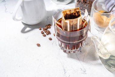 Modaya uygun demlenmiş damlatılmış kahve, soğuk demlenmiş kahve. Damlatma filtresi kahve poşeti cam bardakta beyaz masada, kahverengi şeker, kahve çekirdekleri, sıcak suyla demlik