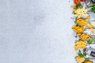 Beyaz masa arkasında çeşitli kurutulmuş spagetti, makarna ve kaşıklarla hazırlanmış makarna. Farklı şekillerde çiğ İtalyan makarnası, otlar, zeytinyağı, domatesler, baharatlar.