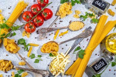 Beyaz masa arkasında çeşitli kurutulmuş spagetti, makarna ve kaşıklarla hazırlanmış makarna. Farklı şekillerde çiğ İtalyan makarnası, otlar, zeytinyağı, domatesler, baharatlar.