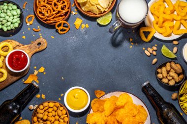 Beer with various salted snacks set. Black table background with traditional party snacks, beer bottles and glasses, with chips, onion rings, salted nuts, crisps and sauces top view copy space