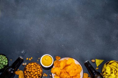 Çeşitli tuzlu abur cuburlarla bira. Siyah masa arkaplanı geleneksel parti atıştırmalıkları, bira şişeleri ve bardaklar, cips, soğan halkaları, tuzlanmış fındık, cips ve soslu üst görünüm fotokopi alanı