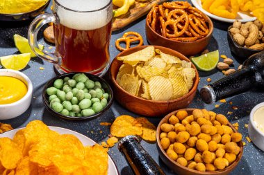 Çeşitli tuzlu abur cuburlarla bira. Siyah masa arkaplanı geleneksel parti atıştırmalıkları, bira şişeleri ve bardaklar, cips, soğan halkaları, tuzlanmış fındık, cips ve soslu üst görünüm fotokopi alanı