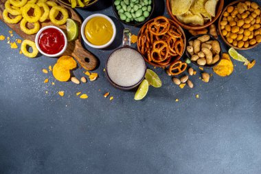 Çeşitli tuzlu abur cuburlarla bira. Siyah masa arkaplanı geleneksel parti atıştırmalıkları, bira şişeleri ve bardaklar, cips, soğan halkaları, tuzlanmış fındık, cips ve soslu üst görünüm fotokopi alanı