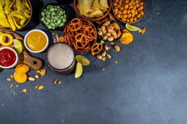 Çeşitli tuzlu abur cuburlarla bira. Siyah masa arkaplanı geleneksel parti atıştırmalıkları, bira şişeleri ve bardaklar, cips, soğan halkaları, tuzlanmış fındık, cips ve soslu üst görünüm fotokopi alanı