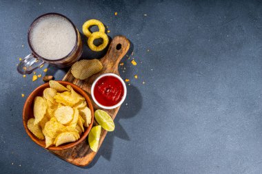 Çeşitli tuzlu abur cuburlarla bira. Siyah masa arkaplanı geleneksel parti atıştırmalıkları, bira şişeleri ve bardaklar, cips, soğan halkaları, tuzlanmış fındık, cips ve soslu üst görünüm fotokopi alanı