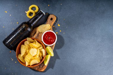 Çeşitli tuzlu abur cuburlarla bira. Siyah masa arkaplanı geleneksel parti atıştırmalıkları, bira şişeleri ve bardaklar, cips, soğan halkaları, tuzlanmış fındık, cips ve soslu üst görünüm fotokopi alanı