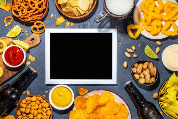 Beer with various salted snacks set. Black table background with traditional party snacks, beer bottles and glasses, with chips, onion rings, salted nuts, crisps and sauces top view copy space