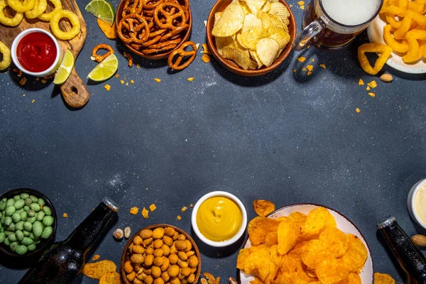 Beer with various salted snacks set. Black table background with traditional party snacks, beer bottles and glasses, with chips, onion rings, salted nuts, crisps and sauces top view copy space