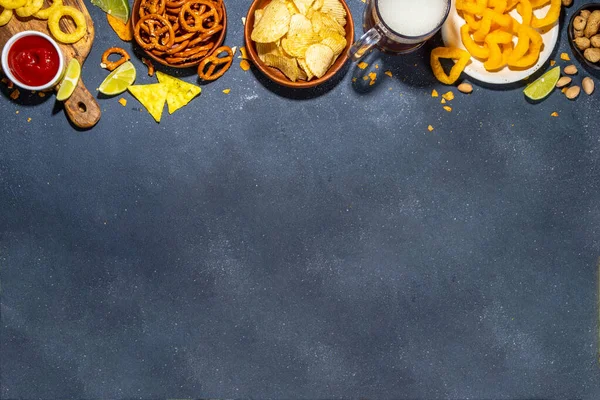 Beer with various salted snacks set. Black table background with traditional party snacks, beer bottles and glasses, with chips, onion rings, salted nuts, crisps and sauces top view copy space