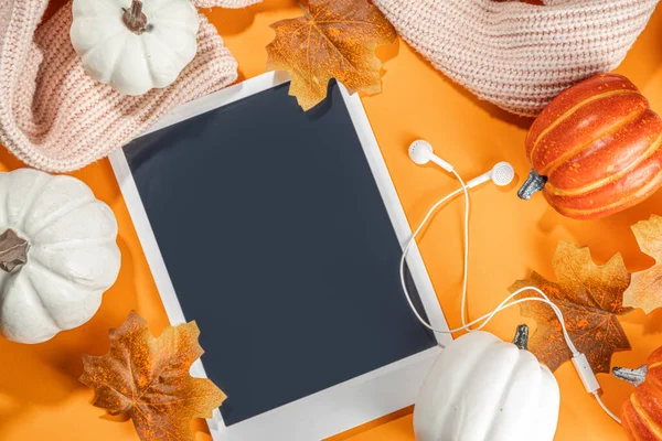 Cozy fall background with tablet, ear-pods, white, orange pumpkins, autumn leaves decor on high-colored orange background. Autumn still life composition.