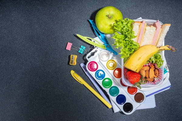 Healthy school lunch box: sandwich, vegetables, fruit, nuts and yogurt with school kids supplies, accessories and backpack on black background flatlay copy space. Back to school concept