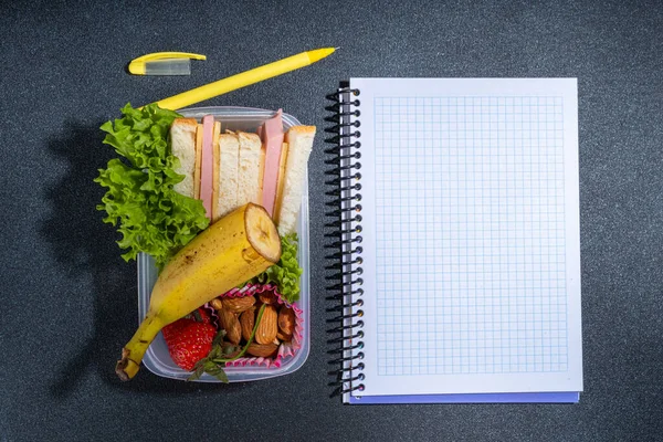 Healthy school lunch box: sandwich, vegetables, fruit, nuts and yogurt with school kids supplies, accessories and backpack on black background flatlay copy space. Back to school concept