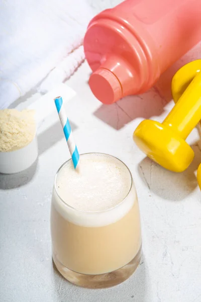 Vanilla white protein shake glass, with straw, with  protein powder, dumbbell, towel and fitness accessories, on white sport bar table background copy space