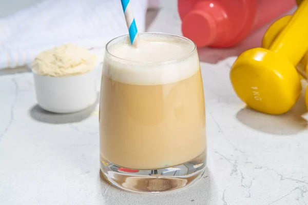 Vanilla white protein shake glass, with straw, with  protein powder, dumbbell, towel and fitness accessories, on white sport bar table background copy space