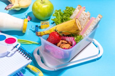 Sağlıklı okul beslenme çantası: sandviç, sebze, meyve, fındık ve yoğurt, içinde okul çocukları malzemeleri, aksesuarlar ve yüksek renkli mavi arka plan fotokopi alanı üzerine sırt çantası