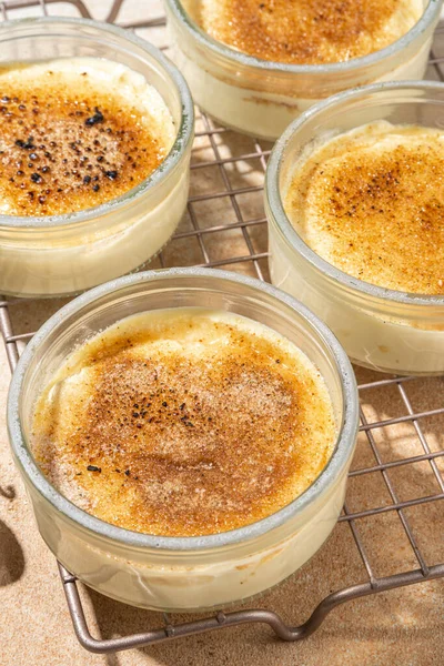 Sweet creme brulee dessert. Portioned glass jars with homemade creme brulee with caramelized sugar crust topping, on beige colored background with sugar in spoon and vanilla copy space