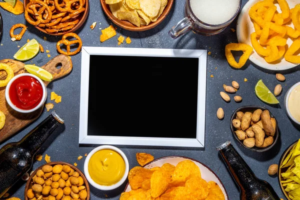 Beer with various salted snacks set. Black table background with traditional party snacks, beer bottles and glasses, with chips, onion rings, salted nuts, crisps and sauces top view copy space