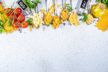 Beyaz masa arkasında çeşitli kurutulmuş spagetti, makarna ve kaşıklarla hazırlanmış makarna. Farklı şekillerde çiğ İtalyan makarnası, otlar, zeytinyağı, domatesler, baharatlar.