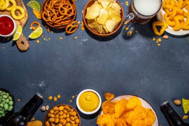 Çeşitli tuzlu abur cuburlarla bira. Siyah masa arkaplanı geleneksel parti atıştırmalıkları, bira şişeleri ve bardaklar, cips, soğan halkaları, tuzlanmış fındık, cips ve soslu üst görünüm fotokopi alanı