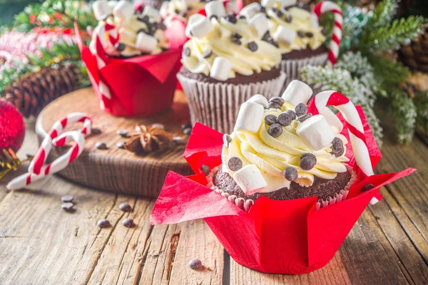 Christmas cupcakes. Xmas hot chocolate cupcake with peppermint candy cane, chocolate drops and marshmallow. Wooden background with Christmas decor and fir tree branches. Sweet Christmas baking/