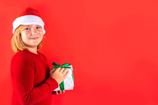 Christmas holiday background with funny blond girl. Little kid girl in red Christmas sweater and funny santa hat costume, with gift box on red background, Xmas greeting card, copy space