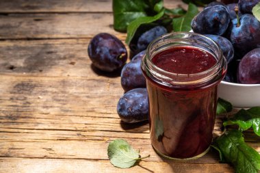 Red blue plum jam in small jar. Homemade autumn plum jam with fresh fruits. Fall preparations and canning on wooden table background copy space