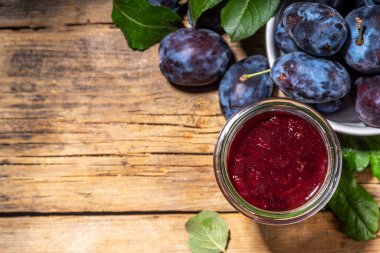 Red blue plum jam in small jar. Homemade autumn plum jam with fresh fruits. Fall preparations and canning on wooden table background copy space
