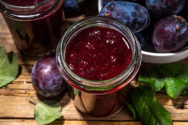 Red blue plum jam in small jar. Homemade autumn plum jam with fresh fruits. Fall preparations and canning on wooden table background copy space