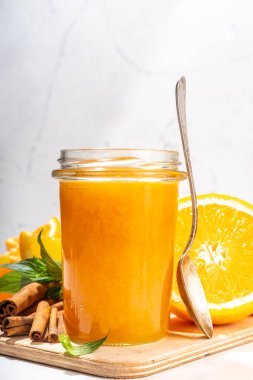 Homemade orange jam in small jar. Homemade orange spicy jam with fresh oranges, cinnamon, mint. Fall preparations and canning on white table background copy space