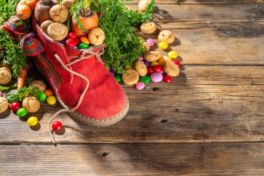 Sinterklaas, Saint Nicholas day background. Little kid red shoe with carrots and sweets, trooigoed, pepernoten, candy. Dutch Christmas holiday on cozy wooden background. 