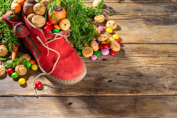Sinterklaas, Saint Nicholas day background. Little kid red shoe with carrots and sweets, trooigoed, pepernoten, candy. Dutch Christmas holiday on cozy wooden background. 