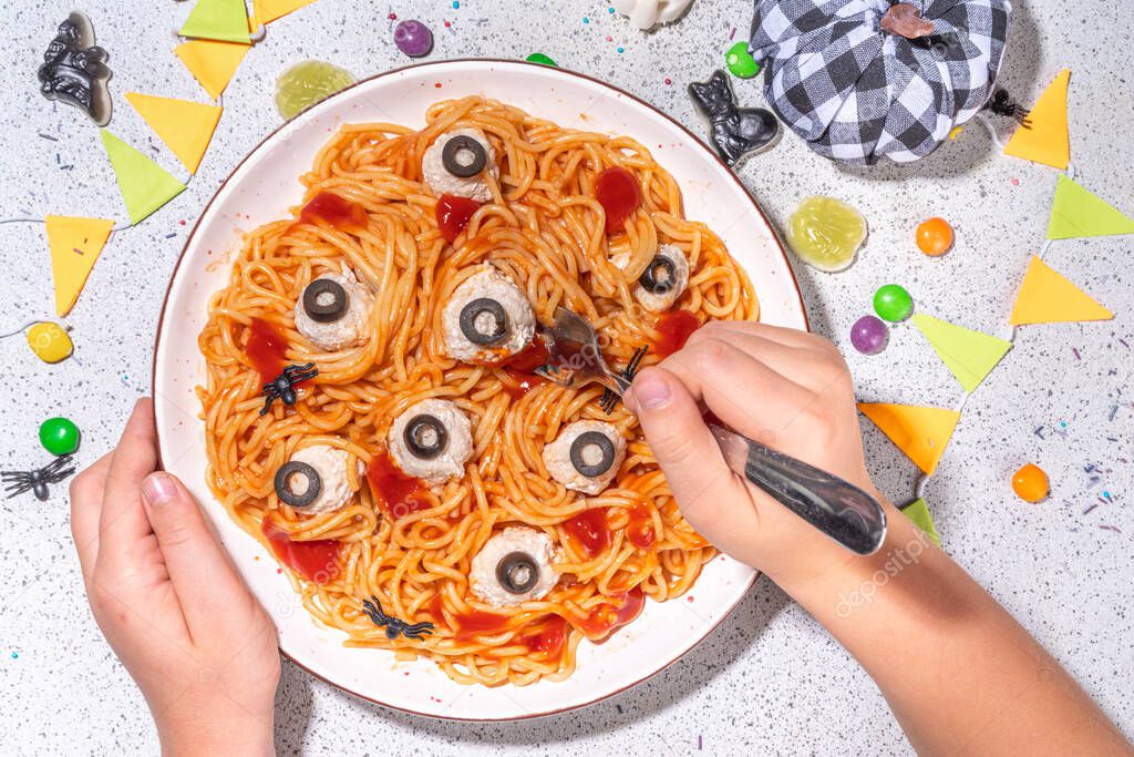 Creepy funny pasta spaghetti for Halloween party dinner. Spaghetti with tomato sauce and chicken meatball decorated with black olives like eyes, white table background with Halloween decorations
