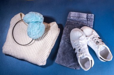 Kadın kış kış sonbahar kıyafetleri seti. Kot pantolon, beyaz örgü süveter ve şapka, koyu mavi arka planda eldivenler ve spor ayakkabılar. Rahat moda kıyafeti düz yatıyordu.