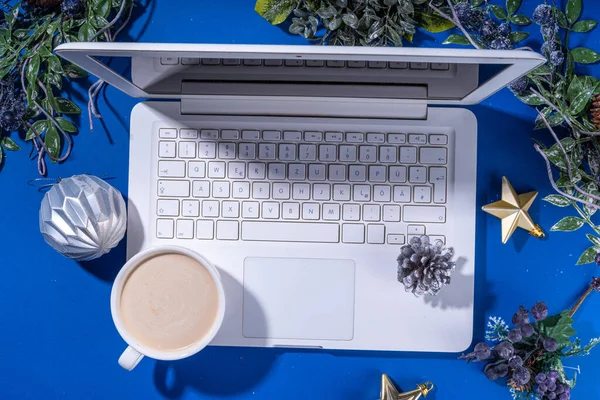 Christmas sale, preparation for holidays concept. Winter office workplace background. Laptop, hot chocolate latte cup with Christmas toys, winter tree branches, flat lay on bright blue background