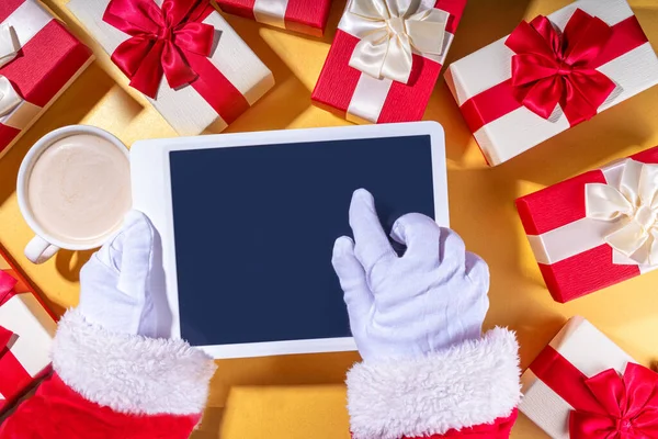 Christmas sale, making wishlist, preparation gifts for Christmas concept. Santa Claus hands with tablet computer, hot chocolate latte cup, gift boxes, flat lay on golden background copy space