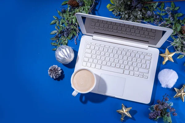 Christmas sale, preparation for holidays concept. Winter office workplace background. Laptop, hot chocolate latte cup with Christmas toys, winter tree branches, flat lay on bright blue background