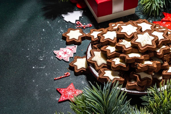 Christmas star sugar glazed cookies, traditional Xmas dark and white chocolate Gingerbread biscuits with holiday decoration, gifts, Christmas tree branch on dark green table top view copy space