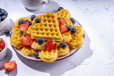Çilekli, yaban mersinli ve ballı Belçika waffle 'ları, beyaz mutfak masalarında ev yapımı kalp şeklinde waffle' lar.