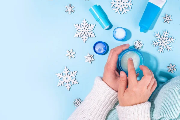 Woman using winter cream for hands. Girl hands with cream flat lay on light blue background with artifical snowflakes and different hand creams. Winter hand skin care cosmetics background
