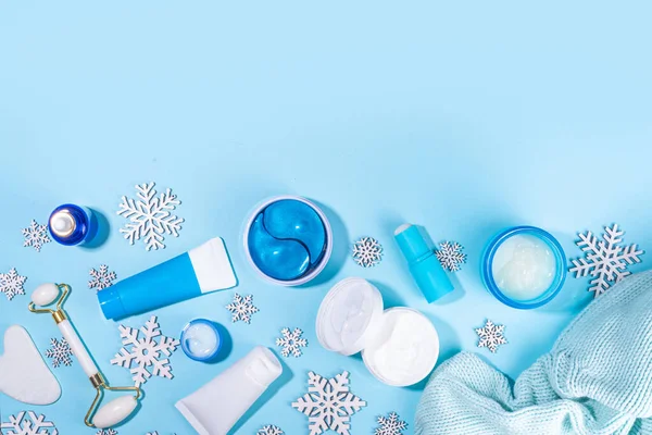 Winter skin care and hand care cosmetic in unbranded containers, bottles, tubes with artificial snowflakes on light blue background. Winter hand skin care cosmetics background top view copy space
