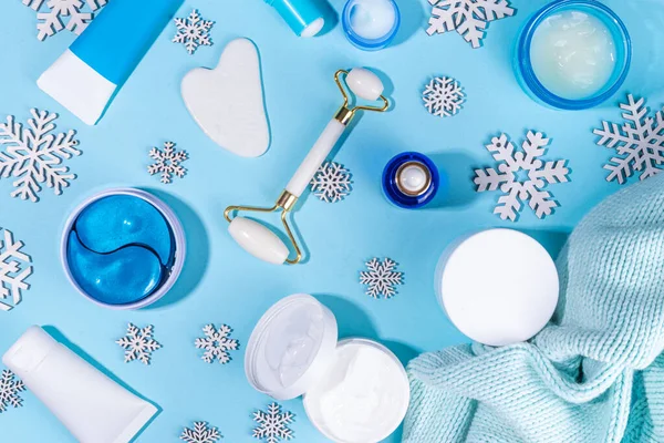 Winter skin care and hand care cosmetic in unbranded containers, bottles, tubes with artificial snowflakes on light blue background. Winter hand skin care cosmetics background top view copy space