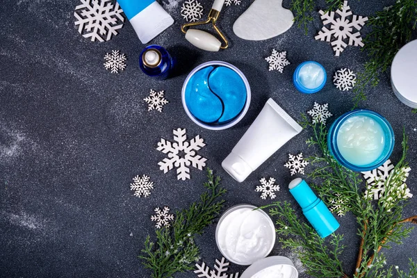 Winter skin care and hand care cosmetic in unbranded containers, bottles, tubes with artificial snowflakes on black stone background. Winter hand skin care cosmetics background top view copy space