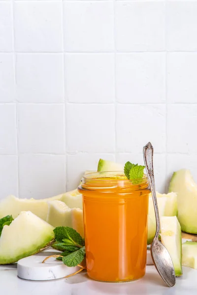 Sweet melon jam, homemade jelly preserves in small glass jar with fresh melon slices on white marble table