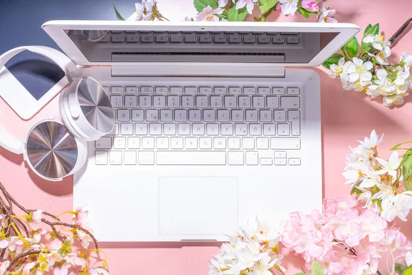 Spring office workplace, blogging flat lay background. White laptop, with headphones, tablet, spring flowers bouquet on pink pearl background, girl\'s hands typing on a laptop.